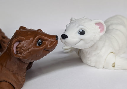 Two 3d printed toy ferrets one is brown and the other is white with pink ears on a soft white background