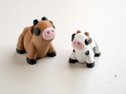 Two toy cow figures, one brown and black, one white with black spots, on a white background.