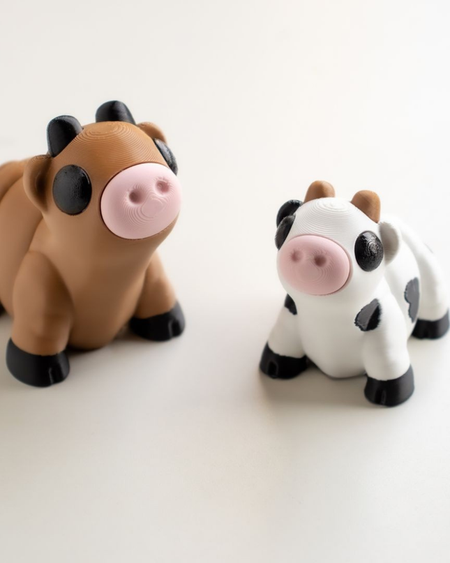 Two toy cow figures, one brown and black, one white with black spots, on a white background.