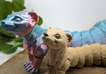 Two 3d printed ferret toys one a shiny rainbow and other is a light tan. standing on a wooden base with a green plant and plain white background