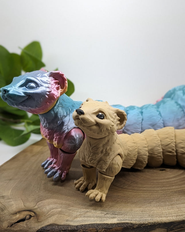 Two 3d printed ferret toys one a shiny rainbow and other is a light tan. standing on a wooden base with a green plant and plain white background