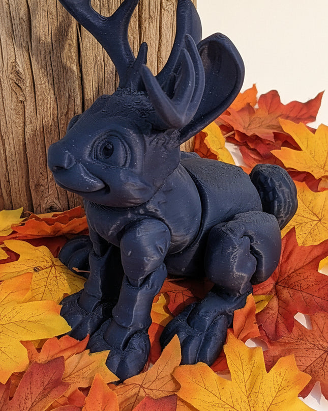 Blue shiny jackalope figurine on a white surface with a wooden stick in the background with colourful maple leaves all around