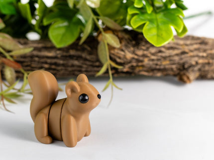 Small brown squirrel figurine on a white surface with green leaves and wood in the background