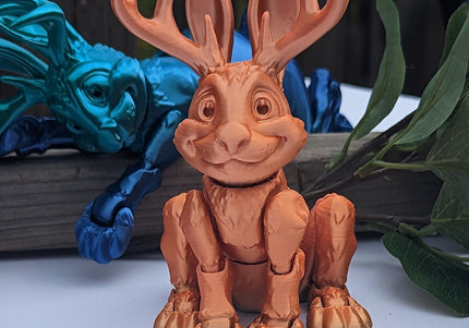 Toy figure of a 3d printed jackalope with a blue one laying in the background with large antlers on a white surface