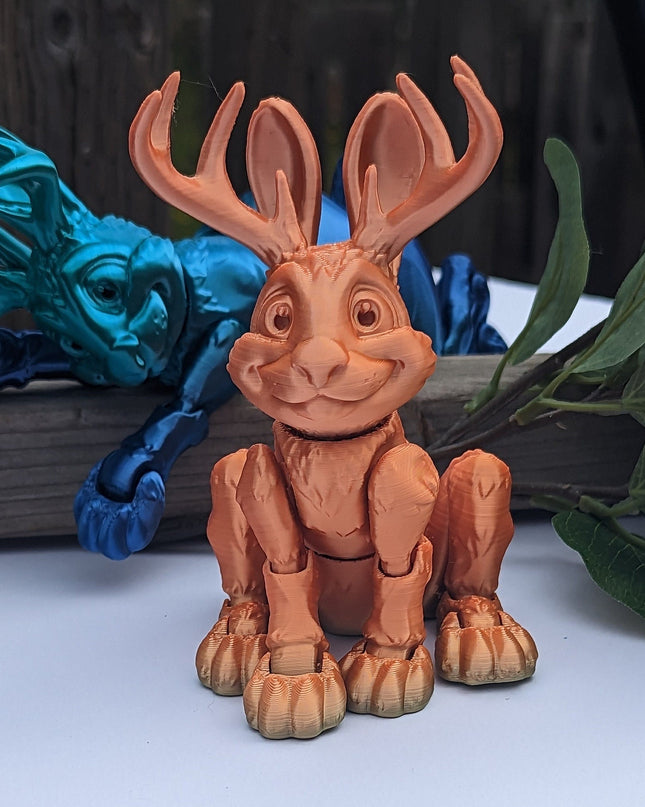 Toy figure of a 3d printed jackalope with a blue one laying in the background with large antlers on a white surface