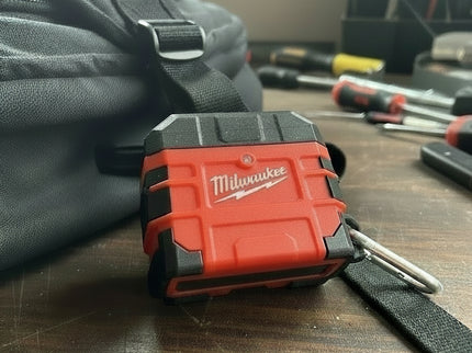 Red and black Milwaukee Air Pod Case closed, on a workbench with a black backpack and tools blurred in the background