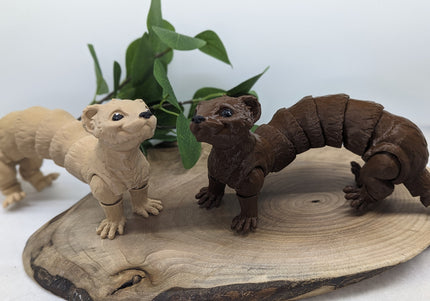 two 3d printed ferret toys one tan/beige the other a chocolate brown on a wooden surface with a green plant in the background