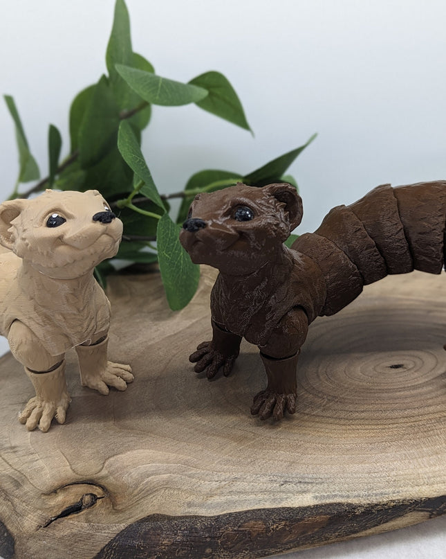 two 3d printed ferret toys one tan/beige the other a chocolate brown on a wooden surface with a green plant in the background