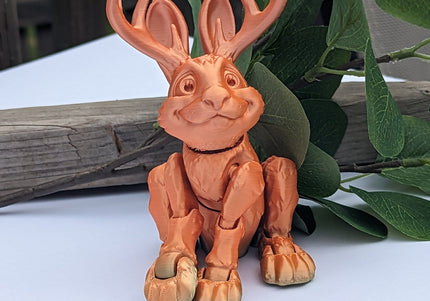 Terracotta-colored figurine of a jackalope with antlers on a white surface with green leaves and wood in the background.