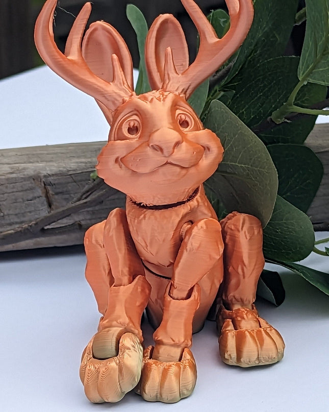 Terracotta-colored figurine of a jackalope with antlers on a white surface with green leaves and wood in the background.