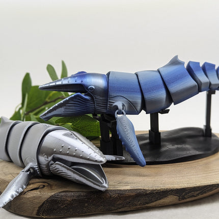 3D-printed model of a humpback whales in a blue ombre in the background with a grey one on a wooden surface with a white background