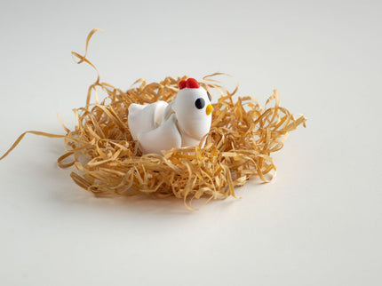 Small white chicken figurine with a red comb in a nest of straw on a light gray background