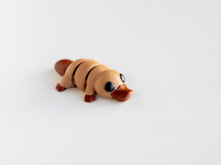 Small toy figure of a platypus which is light and dark brown on a white background