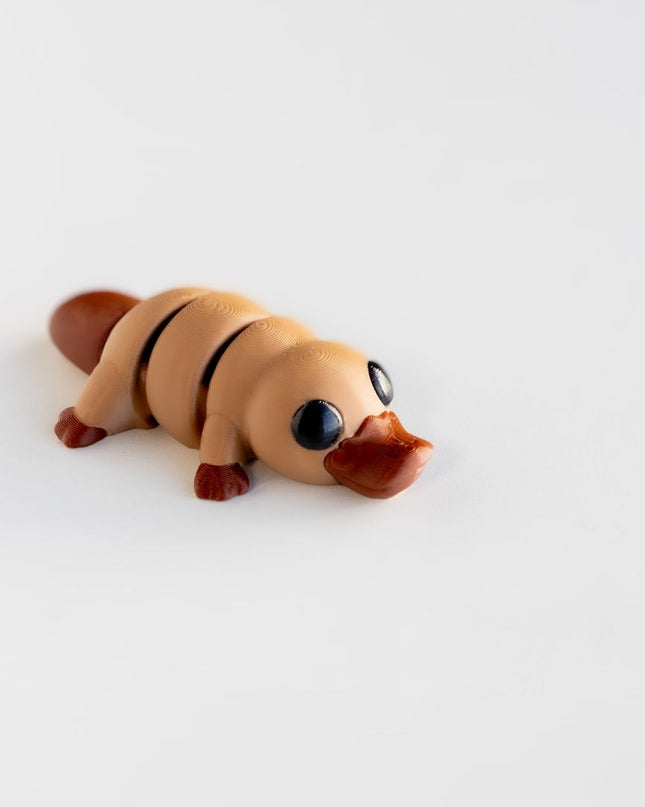 Small toy figure of a platypus which is light and dark brown on a white background