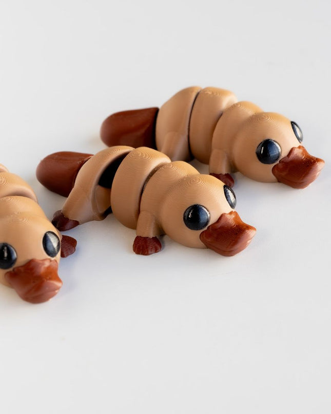 3 platypus articulating toys with a light brown body and dark brown bill and feet, on a white background
