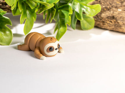 3d printed Toy sloth on a white surface with green leaves and wood in the background