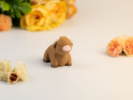 Small brown highland cow toy on a light surface with flowers in the background