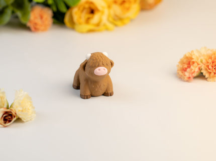 Small brown highland cow toy on a light surface with flowers in the background