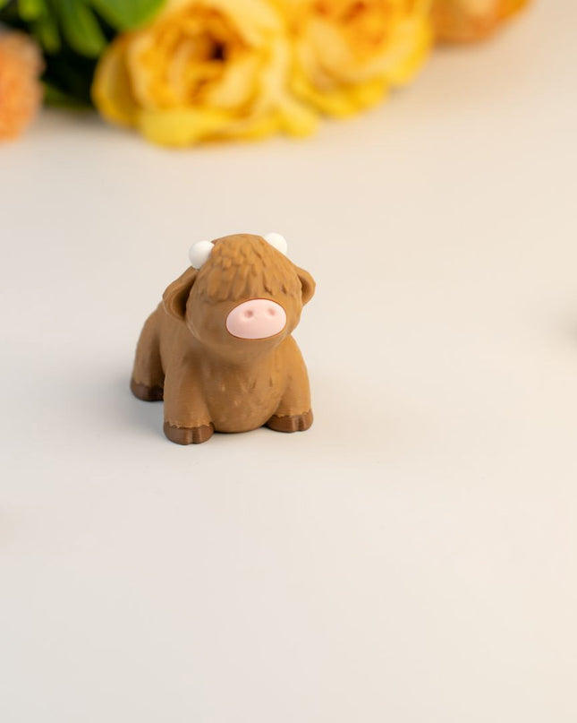 Small brown highland cow toy on a light surface with flowers in the background