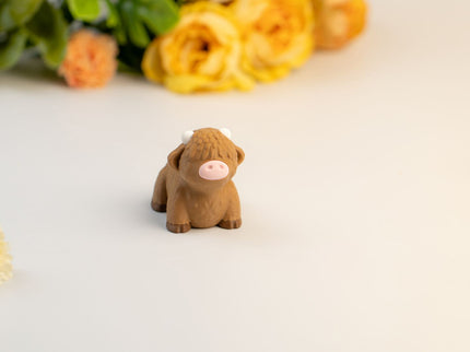 Small brown highland cow toy on a light surface with flowers in the background