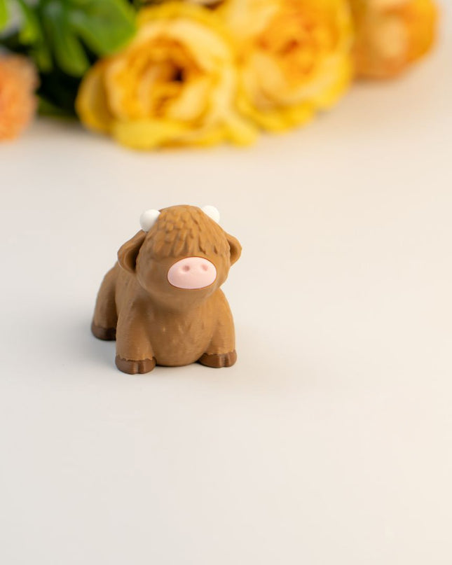 Small brown highland cow toy on a light surface with flowers in the background